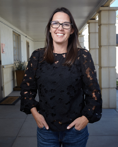 Adrienne Kessler, stands in front of the Speech and Language Development center. She is brunette, wears glasses, and is wearing a black long sleeve blouse. Her hands are in her pockets and she gives the camera a wide and kind smile.