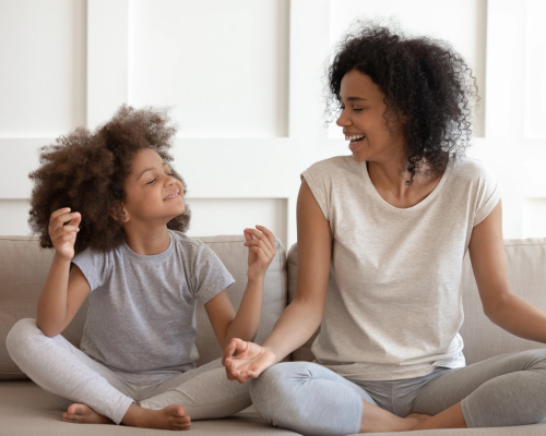 mother teaching daughter how to meditate and practice mindfulness while sitting together on the couch meditating in matching gray outfits