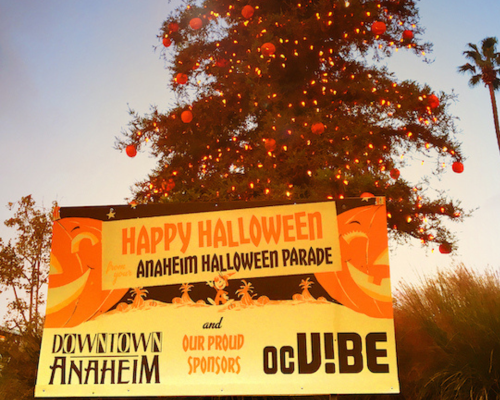 A banner with Anaheim Fall Festival and Halloween Parade in front of a big tree with lights